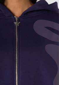 Felpa con zip colore navy, dotata di una cerniera argento e un tirante a forma di farfalla. Presenta un grande design astratto di colore viola chiaro sulla parte anteriore. Tessuto morbido.