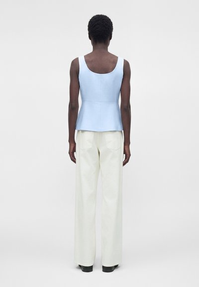 Άτομο με κοντά μαλλιά που φοράει ανοιχτό μπλε αμάνικη μπλούζα και λευκό παντελόνι με φαρδιά πόδια, στέκεται πλάτη σε ένα απλό γκρι φόντο.