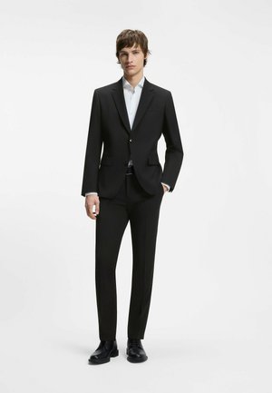 Joven de pie vistiendo un traje negro a medida con camisa blanca y zapatos de vestir negros sobre un fondo blanco liso.
