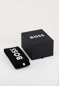 Svart eske med "BOSS" i hvitt på toppen, ved siden av en svart fløyelspose med det samme logoen. Posene har et lite tagginnehold.