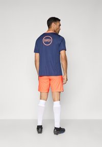 Mornarska sportska majica s "BARÇA" logotipom na leđima, uparen sa svijetlo narančastim šorcama. Bijele čarape do koljena i crne nogometne kopačke.