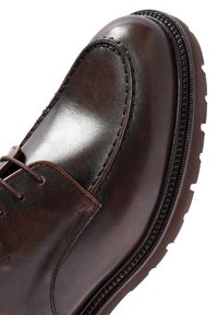 Zapato de cuero marrón con un acabado brillante, que presenta una punta redonda, detalles cosidos y una suela texturizada con estrías para mayor agarre.