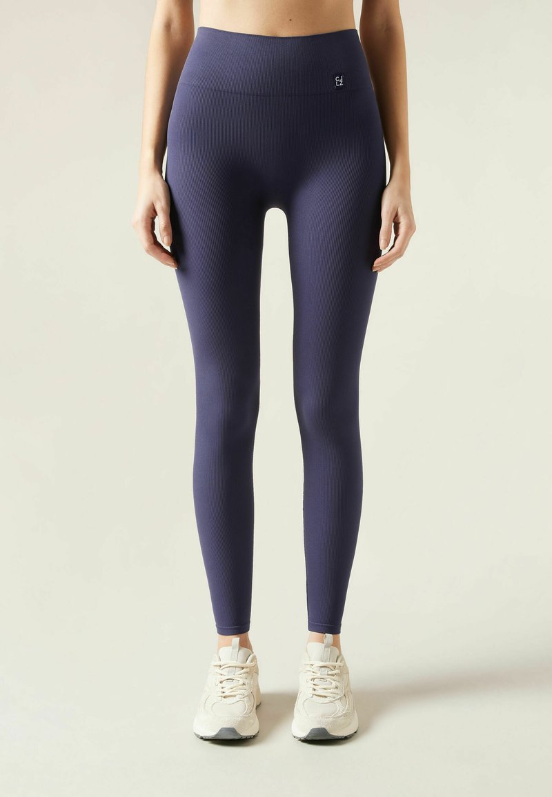 Leggings morados con relieve y cintura alta, textura suave y detalle sutil del logo, combinados con zapatillas deportivas blancas.