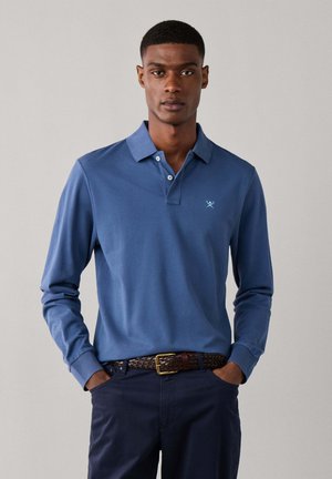 Joven con una camisa polo azul de manga larga con un pequeño logo y pantalones oscuros, de pie con las manos en los bolsillos sobre un fondo liso.