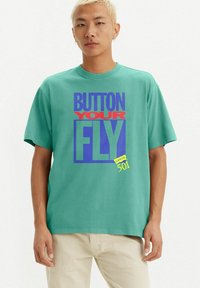 Turquoise cotton t-shirt featuring a graphic design with the text "BUTTON YOUR FLY" in red, blue, and yellow. Short sleeves and crew neck.