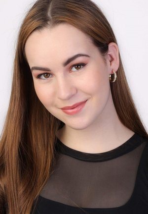 Portret van een persoon met lang bruin haar, die gouden hoepeloorbellen draagt, een zwarte top met een transparante halslijn en een neutrale make-uplook.