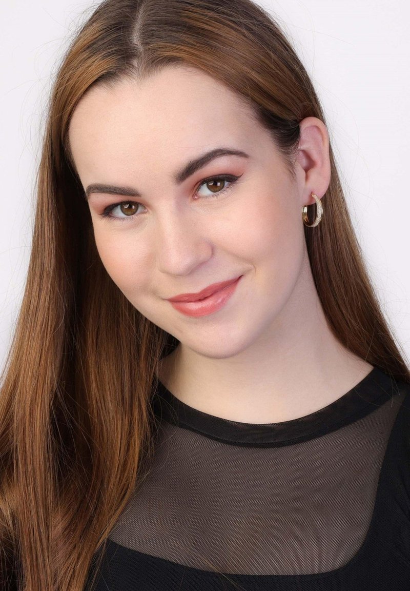 Portrait d'une personne avec de longs cheveux bruns, portant des boucles d'oreilles en or, un haut noir avec un décolleté transparent, et un maquillage neutre.