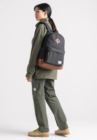 Zaino nero con sezione inferiore marrone, dettaglio a forma di diamante e tasca frontale. Persona vestita con un outfit verde oliva sta in posizione laterale.