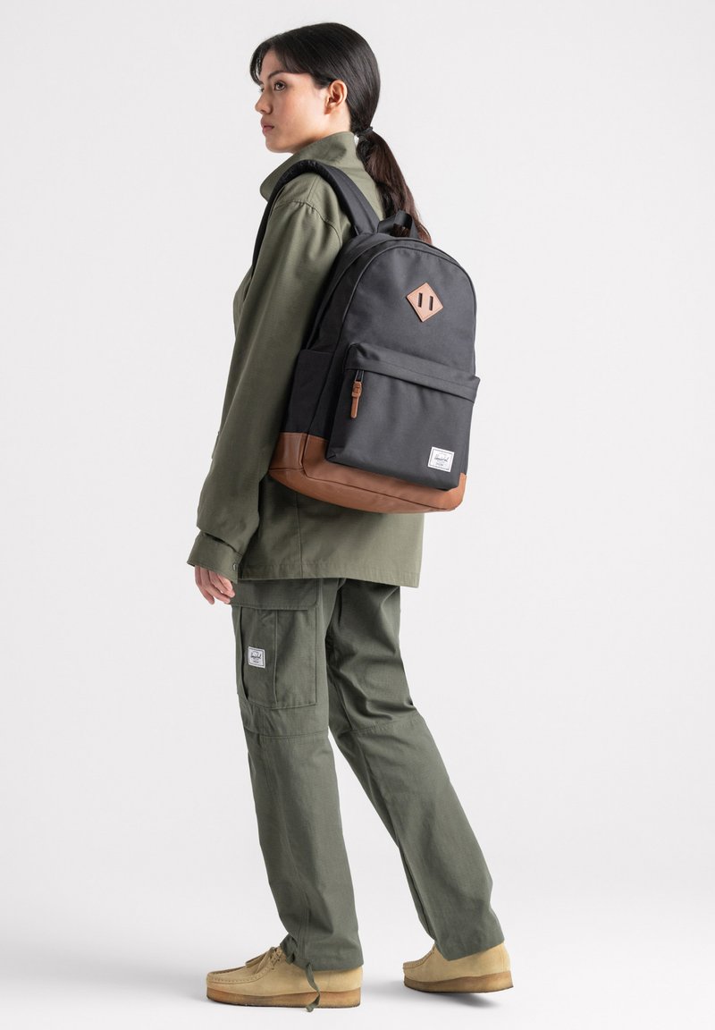 Zaino nero con sezione inferiore marrone, dettaglio a forma di diamante e tasca frontale. Persona vestita con un outfit verde oliva sta in posizione laterale.