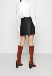 Mini-jupe en cuir noir avec finition texturée, assortie à une chemise blanche à manches longues et des bottes en cuir marron jusqu'aux genoux avec des talons carrés.