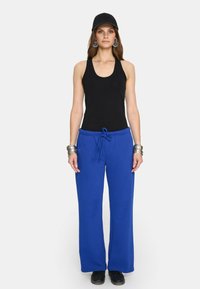 Kobieta w czarnej bluzce bez rękawów, czapce w czarnym kolorze, niebieskich spodniach ze sznurkiem, dużych srebrnych kolczykach oraz srebrnych bransoletkach, stojąca na tle białym.