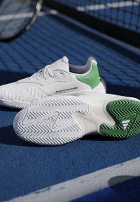 Sneakers bianche e verdi con suole strutturate posizionate su un campo da tennis blu vicino a una rete.