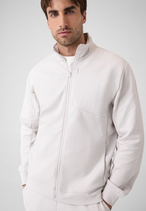 Mann mit kurzen braunen Haaren trägt eine hellgraue Reißverschlussjacke mit hohem Kragen und steht mit den Händen in den Taschen vor einem schlichten Hintergrund.