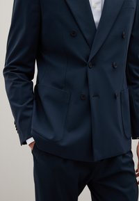 Blazer doppiopetto blu navy con ampie tasche a pattina, texture liscia e bottoni neri. Design aderente con maniche su misura e rever.