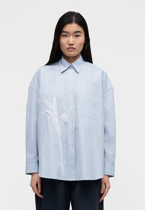 Žena nosící volnou světle modrou pruhovanou košili na knoflíky s bílým vyšívaným motivem rostliny na levé straně, v kombinaci s tmavými kalhotami.