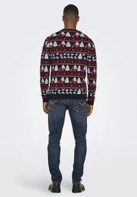 Maglione natalizio nero con motivi rossi e bianchi che presentano alberi e fiocchi di neve, abbinato a jeans blu e stivali neri.