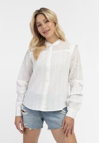 Witte langarmshirt met verticale strepen, een hoge kantenkraag en oogjesdetails op de mouwen. Gepaard met versleten denimshorts.