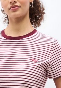 Γυναίκα που φοράει μαύρο-κόκκινη ριγέ μπλούζα Levi's με κρίκους σκουλαρίκια, τα σγουρά μαλλιά της είναι πιασμένα πίσω, ορατή από τον λαιμό μέχρι τη μέση.