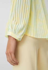 Blusa gialla plissettata con motivo di foglie verde chiaro, maniche lunghe e polsini elastici, abbinata a pantaloni attillati giallo chiaro.