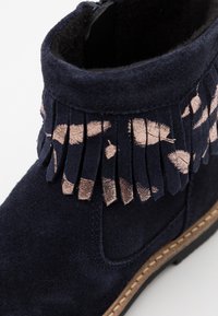 Námořnické semišové kotníkové boty s fringingovým designem a metalickými růžovými zlatými akcenty a béžovou podrážkou. Hladký povrch, kulatý tvar špičky.