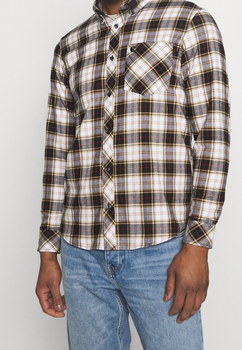 Camicia a scacchi in bianco, nero e tonalità di marrone, con tasca sul petto, maniche lunghe e orlo curvo. Indossata con jeans blu.