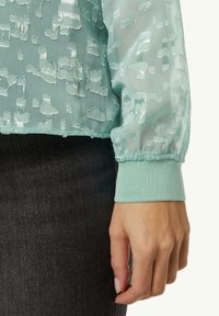 Langärmlige, mintgrüne Bluse mit Textur und transparenter Optik, kombiniert mit schwarzen Jeans für einen Kontrast.