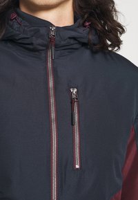 Tmavě modrá bunda na zip s kapucí, se burgundskými akcenty, strukturovanou látkou a dvěma kapsami na zip na přední straně.