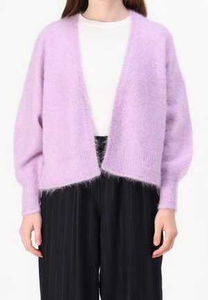 Personne portant un cardigan lavande duveteux ouvert sur le devant, par-dessus un haut blanc et un pantalon large noir à fines rayures, sur un fond blanc.