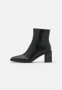 Bottine en cuir noir avec un bout pointu, une texture lisse et un talon carré épais, présentant un design sans couture et un matériel minimal.