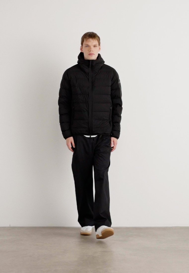 Veste noire matelassée avec col montant, fermeture éclair et poches latérales ; assortie à un pantalon noir ample et des baskets blanches.