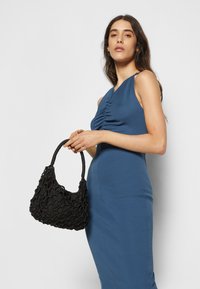 Vestido justo azul com franzidos laterais, combinado com uma bolsa preta texturizada com um design em nó e uma alça trançada.