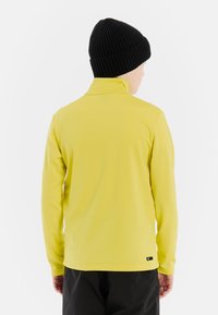 Langsleeve gele shirt met een verhoogde kraag, gemaakt van een gladde stof. Draagt een zwarte geribbelde beanie. Zwarte merklabel zichtbaar aan de zoom.