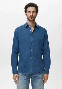 Mann mit kurzen lockigen Haaren und Bart, der ein knöpfbares blaues Jeanshemd und hellblaue Jeans vor einem schlichten Hintergrund trägt.