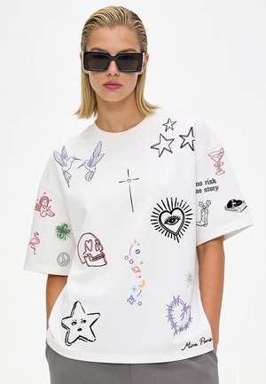 Femme aux cheveux blonds et lunettes de soleil rectangulaires noires porte un t-shirt blanc oversize avec des gribouillis graphiques colorés et des symboles.