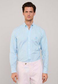 Camisa de botones azul claro con rayas verticales en azul más oscuro. Presenta un pequeño logo en el pecho y puños abotonados. Textura de tela suave.