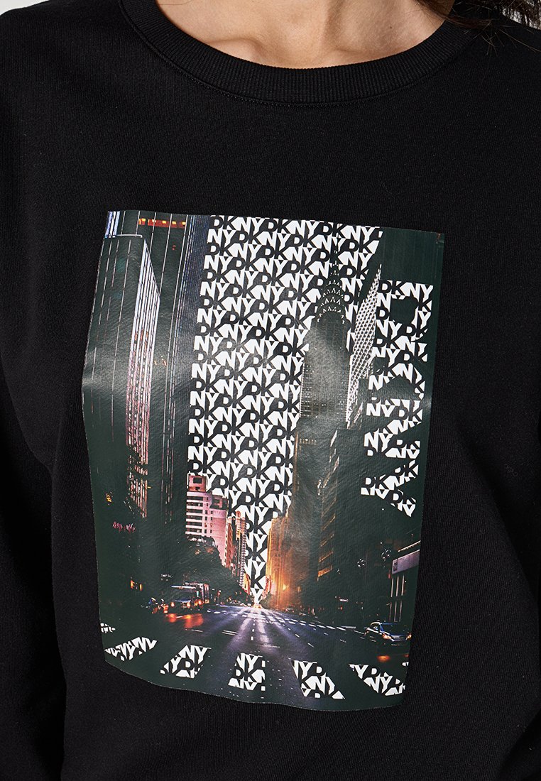 Sudadera negra con un gráfico central de una calle de la ciudad, superpuesto con texto repetido "DKNY" en blanco y formas audaces.