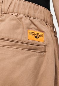 Beige broek met elastische tailleband, achterzak met biezen, en een klein oranje rechthoekig label met de tekst "Service Works".