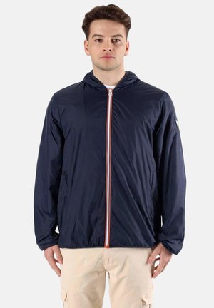 Jeune homme portant une veste zippée à capuche bleu marine et un pantalon cargo beige, debout devant un fond uni gris clair.