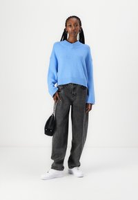 Lys blå strikket genser med v-hals, kombinert med løse mørkegrå jeans og hvite sneakers. Tilgjengelig svart crossover-veske.