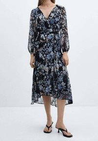 Vestido envolvente floral en negro con flores azules y durazno. Mangas largas translúcidas, escote en V y dobladillo asimétrico. Combinado con sandalias negras de tacón.