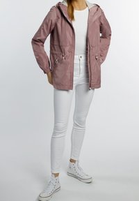 Jasnoróżowa, wodoodporna kurtka z regulowanym paskiem w talii, kapturem i elastycznymi mankietami; w zestawie z białymi dopasowanymi dżinsami i białymi sneakersami.