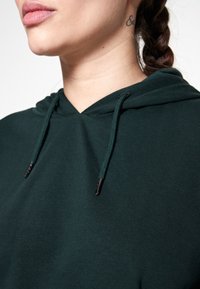 Sudadera verde oscura con cordón con puntas de metal, textura de tela suave y escote frontal visible en el primer plano.