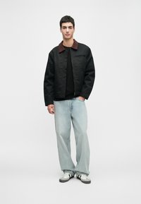 Jaunas vyras stovi, apsirengęs juodu švarku su rudais apykaklėmis, juoda marškinėliais, šviesiai mėlynomis plačiomis džinsomis ir baltais sportbačiais su juodomis juostelėmis.