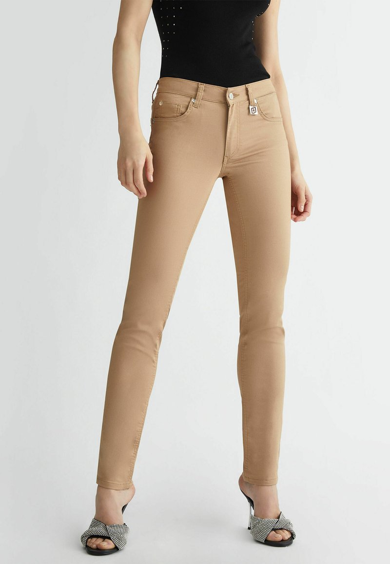 Jeans skinny beige con una texture liscia, cinque tasche e chiusura con bottone. Indossati con sandali neri con tacco e una fascia testurizzata.