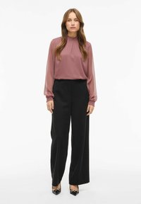 Blusa de gasa color malva con cuello recogido, combinada con pantalones negros de tiro alto. Los pantalones son de pierna ancha y los zapatos tienen un detalle dorado.