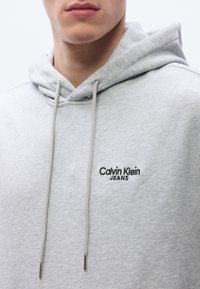 Γκρι φούτερ με κουκούλα, κατασκευασμένο από μαλακό ύφασμα, με κορδόνι στην κουκούλα και μαύρο κέντημα με το λογότυπο "Calvin Klein JEANS" στο στήθος.