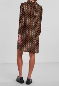 Brun mönstrad klänning med vågiga mönster i rött, gult och svart. Har en fluga vid halsen och långa ärmar, tillsammans med svarta skor.