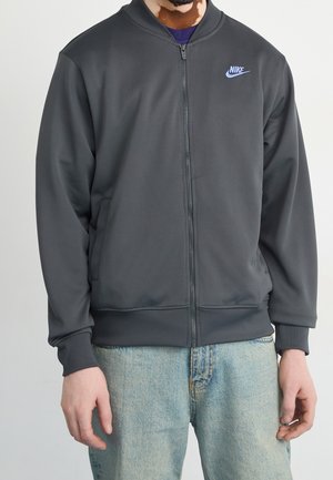 Man draagt een donkergrijze Nike-jas met volledige rits en zakken en lichtblauwe jeans, staand tegen een effen achtergrond.