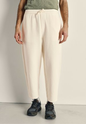 Pantalons joggers beige clair en tissu texturé avec une ceinture élastique et un cordon de serrage. Portés avec des chaussures de sport foncées.