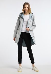 Jachetă lungă gri cu glugă, având accente de culoare albastru deschis și închidere cu fermoar, purtată peste o cămașă albă și pantaloni negri. Textură netedă.